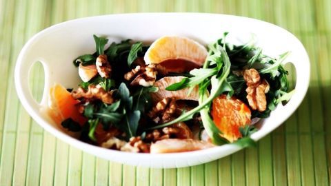 Ein Salat aus Rucola und Mandarinen wird mit Walnüssen in einer weißen Schüssel angerichtet und auf einem grünen Untergrund präsentiert.