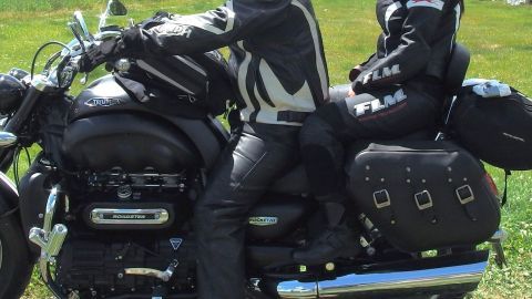 Zwei Personen in Motorradbekleidung aus Leder fahren auf einem Motorrad entlang einer grünen Wiese.