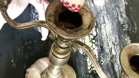 Ein Edelstahl-Kerzenständer wird mit einem Tuch gereinigt, während Wachsreste in einer Umgebung mit Farbspritzern liegen.
