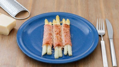 Gratinierter Spargel liegt auf einem blauen Teller, umgeben von Besteck, während Käse gerieben wird.
