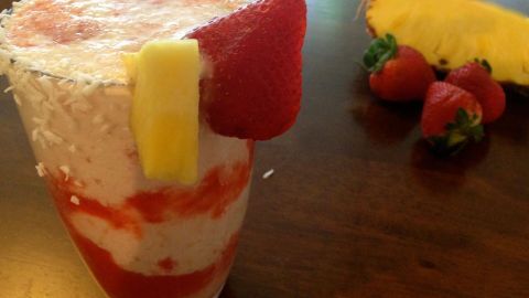 Ein erfrischender Erdbeer-Kokos-Buttermilch Drink in einem Glas, garniert mit Erdbeeren und Ananasstücken auf einem Holztisch.