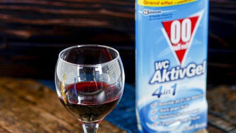 Rotweinflecken auf hellen Kunstfasertextilien mithilfe von WC-Ente erfolgreich entfernen. Ein Glas Rotwein steht auf einem Holztisch, während nebenan eine Flasche WC-Ente AktivGel zu sehen ist.