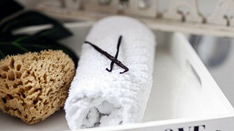 Eine Vanilleschote liegt auf einem gewickelten Handtuch in einem Badezimmer-Setup mit einem Schwamm und dekorativen Elementen.