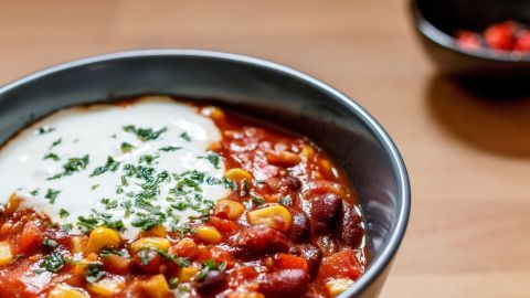 Eine Schüssel Chili sin Carne ist gefüllt mit bunten Bohnen, Gemüse und einem Klecks veganer Creme, umgeben von einem Holztisch.