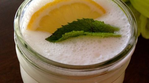 Ein cremiger Zitronen-Buttermilch-Smoothie in einem Glas mit Zitronenscheibe und Minze, auf einem dunklen Tisch.