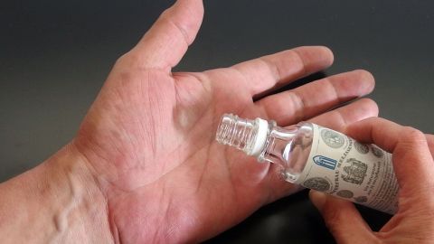Mit Melissengeist Sekundenkleber von Händen entfernen. Eine Hand hält eine Flasche Klosterfrau Melissengeist, während sie auf die andere Hand mit Sekundenkleber zeigt.