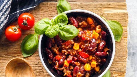 Eine Schüssel Chili con Carne ohne Fix steht auf einem Holzbrett, umgeben von frischem Basilikum und Tomaten.