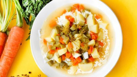Eine Schüssel Salzgurken-Gemüsesuppe mit buntem Gemüse und frischen Kräutern auf einem gelben Tisch, umgeben von Karotten.