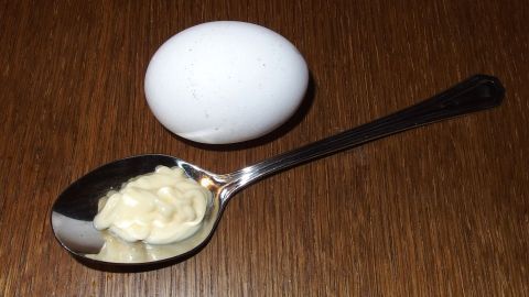Ein Löffel mit Mayonnaise steht neben einem rohen Ei auf einem Holzuntergrund, bereit für eine Haarkur.