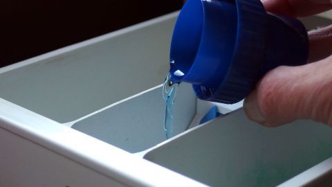 Eine Hand gießt flüssiges Waschmittel aus einem blauen Behälter in ein Waschmittelfach zum Vermeiden von muffiger Wäsche.