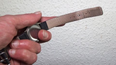 Ein handgehaltenes Lederarmband wird überprüft, um Gerüche mit Apfelessig zu entfernen, vor einer neutralen Wand.