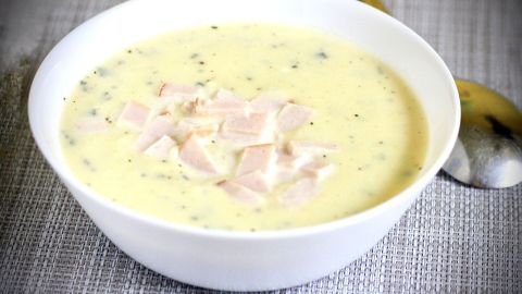 Eine schlichte weiße Schüssel enthält eine cremige KaPü-Suppe mit kleinen, geschnittenen Einlagen. Sie steht auf einem strukturierten, hellen Tisch.