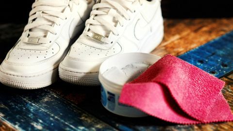 Weiße Turnschuhe liegen neben einer Reinigungscreme und einem pinken Tuch auf einem hölzernen Boden.