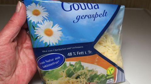 Reibekäse bleibt im Tiefkühlfach ewig lange haltbar. Eine Hand hält eine Verpackung mit Reibkäse, auf der „Gouda geraspelt“ steht, vor einem hellen Hintergrund.
