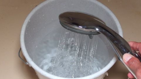 Duschen mit Durchlauferhitzer: Wasser vor dem Duschen auffangen und zum Gießen der Blumen weiterverwenden. Eine Hand hält eine Duschbrause, die Wasser in einen Eimer mit klarem Wasser sprüht, vorbereitet fürs Duschen.