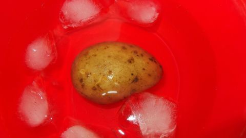Kartoffeln nach dem Kochen 5 Sekunden in Eiswasser baden und schon lässt sich die Schale ohne Probleme abziehen. Eine Kartoffel liegt in einer roten Schüssel mit Eiswasser, um das Schälen zu erleichtern und schneller zu machen.