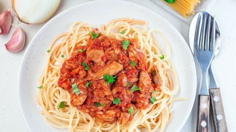 Eine Portion Spaghetti mit Thunfischsoße liegt auf einem Teller, umgeben von Zutaten wie Zwiebel und Knoblauch.