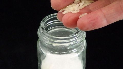 Eine Hand streut fein gemahlenes Salz aus einem Glas auf eine leichte Oberfläche, während der dunkle Hintergrund scharf kontrastiert.