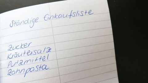 Ein Notizbuch mit der Überschrift „Ständige Einkaufsliste“ zeigt eine Liste mit Lebensmitteln wie Zucker und Zahnpasta.