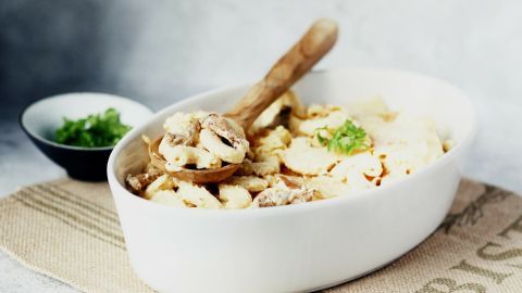 Ein weißes Geschirr mit überbackenem Gyros wird mit einem Holzlöffel serviert, umgeben von frischem Grün.