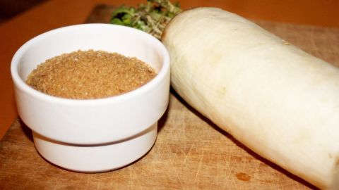 Ganz ohne Chemiebomben wird man den Schleim im Hals mit einem selbsgemachten Saft aus Rettich und Zucker los. Ein Rettich liegt neben einer weißen Schüssel mit Zucker auf einem Holzbrett, bereit zur Zubereitung eines Hustenlöser.