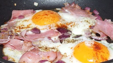 In einer Pfanne braten Schinken und zwei Spiegeleier, umgeben von Zwiebeln, für das Gericht Strammer Max.