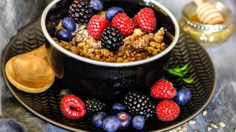 Ein schwarzer Schüssel gefüllt mit knusprigem Müsli, garniert mit frischen Beeren und einem Holzlöffel, liegt auf einem Teller.