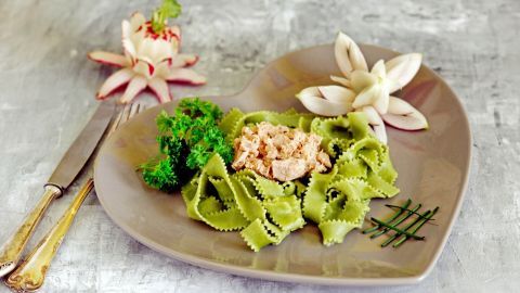 Grüne Nudeln mit Thunfisch-Sahne-Soße liegen auf einem herzförmigen Teller, umgeben von frischen Kräutern und dekorativen Blumen.