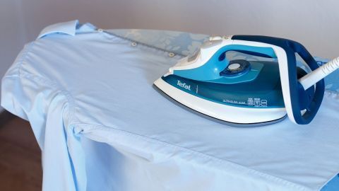 Ein blaues Bügeleisen marke Tefal glättet ein hellblaues Hemd auf einem Bügelbrett in einem hellen Raum.