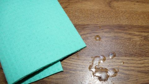 Ein grünes Tuch liegt neben einem Wachsfleck auf einer Holzoberfläche, bereit zum Entfernen der Flecken.