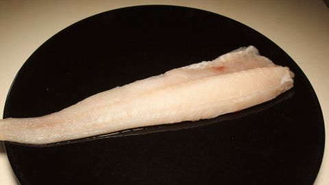 Seelachs (Köhler), ein naher Verwandter von Kabeljau und Schellfisch, kostet meist nur halb so viel wie der Kabeljau, und schmeckt auch sehr gut. Ein Fischfilet liegt auf einem schwarzen Teller, bereit zum Kochen und serviert in einer modernen Küchenszene.