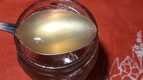 Ein Löffel mit transparentem Akazien- und Holunderblütengelee schimmert im Glas auf einem roten Tisch.