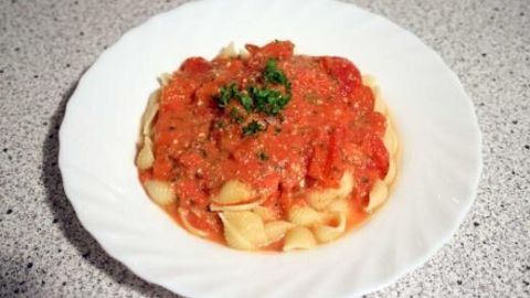 Eine Schüssel Nudeln mit Tomatensauce und Kräutern vor einem hellen, strukturierten Hintergrund.