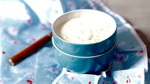 Ein blauer Schüssel mit geronnener Sahne steht auf einem blauen, floral gemusterten Untergrund neben einem Holzlöffel.