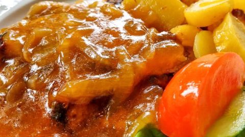 Leckere Zwiebelsoße bedeckt ein saftiges Gericht, umgeben von Kartoffeln und einem frischen Tomatenstück auf Salat.