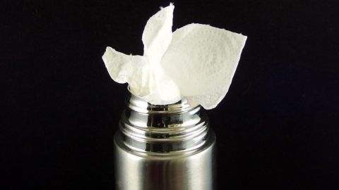 Thermoskanne im Büro reinigen, mit Küchenpapier und heißem Wasser. Eine silberne Thermoskanne hat ein Stück Küchenpapier, das im Öffnungsbereich steckt, um die Reinigung im Büro zu unterstützen.