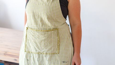 Vermeide Flecken in der Küche, indem du eine praktische Schürze trägst. Die Schürze schützt deine Kleidung beim Kochen und reduziert das Risiko von hartnäckigen Flecken. So bleibt dein Lieblingsoutfit sauber! Eine Person trägt eine karierte Schürze in einer Küche, um Flecken auf der Kleidung zu vermeiden.