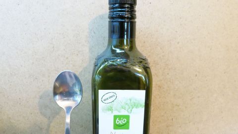 Eine Flasche Olivenöl steht neben einem Löffel auf einer neutralen Oberfläche, bereit zum Verzehr bei Halskratzen.