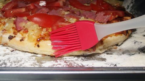 Wenn man nach 5-10 Minuten den Ofen öffnet und mit einem Pinsel den Rand der Pizza mit Speiseöl bestreicht, dann ist der Rand nach weiteren 10 Minuten lecker kross & knusprig. Ein knuspriger Pizzarand wird mit einem roten Pinsel bestrichen, während er auf einem Blech im Ofen liegt.