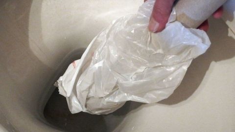 Eine Hand hält eine gefaltete Plastik-Tüte über die Toilette, während die Toilette verstopft ist, ohne Saugglocke zur Hand.