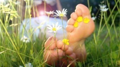 Füße liegen entspannt im Gras, umgeben von Gänseblümchen und Sonnenlicht, mit gelben Blütenblättern zwischen den Zehen.