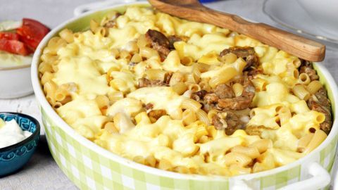 Ein Auflauf mit Gyros, Makkaroni und geschmolzenem Käse wird in einer grünen Auflaufform serviert, umgeben von Beilagen.
