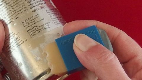 Ein blauer Radiergummi entfernt Aufkleberreste von einem Glasbehälter vor rotem Hintergrund.