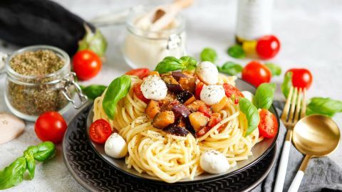 Pasta mit Auberginen und Tomaten ist appetitlich angerichtet, umgeben von frischen Kräutern und Kirschtomaten auf einem Teller.