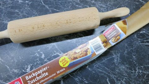 Nudelholz liegt still neben einer geöffneten Packung "Backpapier-Zuschnitte" auf einer dunklen, marmorierten Fläche.
