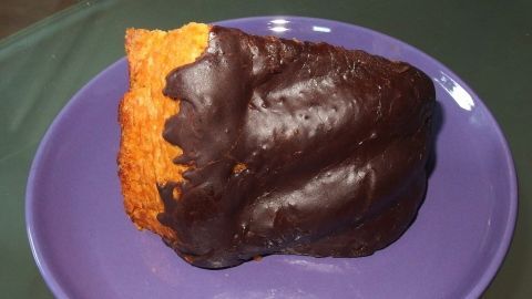 Ein Stück Kuchen mit selbstgemachter Schokoglasur liegt auf einem lila Teller, auf einem grünen Tisch.