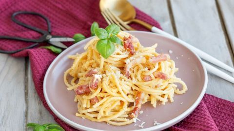 Ein Teller mit "echter" Carbonara, gekrönt mit frischem Basilikum, auf einem Tisch mit violettem Tuch und Besteck.
