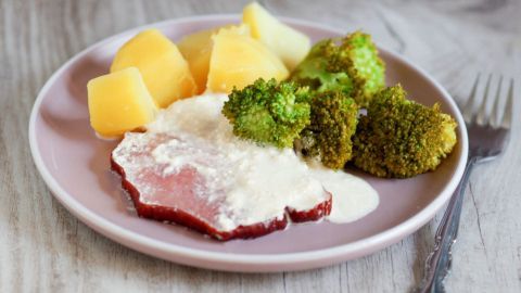 Eine Portion Kasseler liegt auf einem Teller, umgeben von Schmandsoße, Kartoffeln und Brokkoli auf hellem Holz.