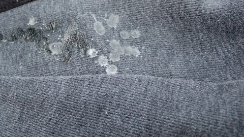 Ein graues Kleidungsstück liegt auf einer Oberfläche, mit mehreren Wachsflecken, die entfernt werden sollen, möglicherweise mit Orangenöl.