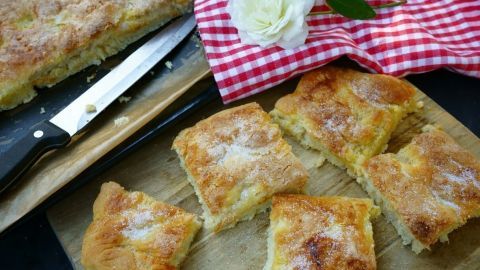 Zuckerkuchen aus Hefeteig liegt auf einem Holzbrett, in Stücke geschnitten, mit einer Rose und kariertem Tuch im Hintergrund.